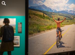Der Blick in die Ausstellung zeigt das großformatige Foto eines Fahrradfahrers in den Bergen, im Hintergrund betrachtet eine Frau weitere Bilder