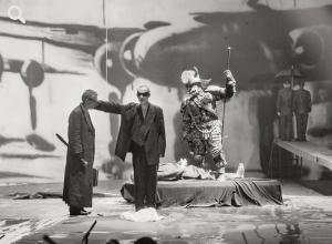 Foto einer Theaterszene mit mehreren Männern, einer davon in einem monsterartigen Kostüm