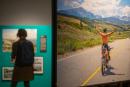 Der Blick in die Ausstellung zeigt das großformatige Foto eines Fahrradfahrers in den Bergen, im Hintergrund betrachtet eine Frau weitere Bilder